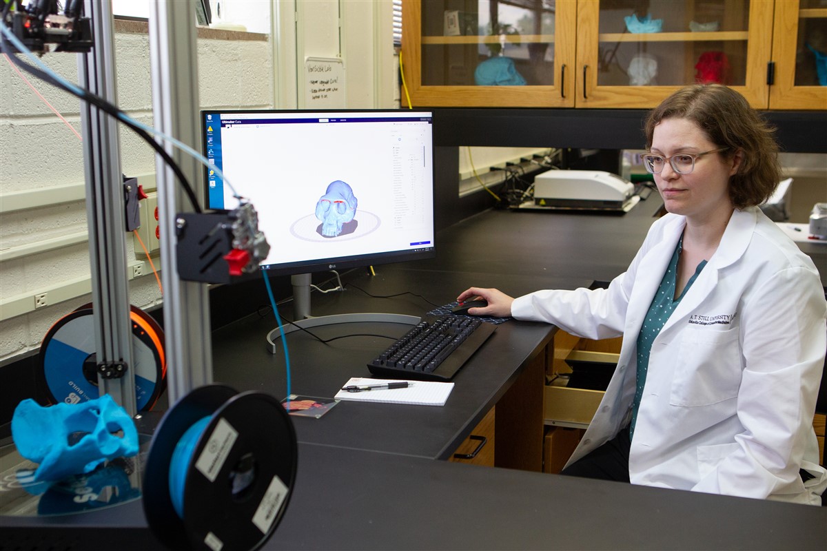 Woman uses a computer in a lab while watching a 3D printer print a fossil pelvis